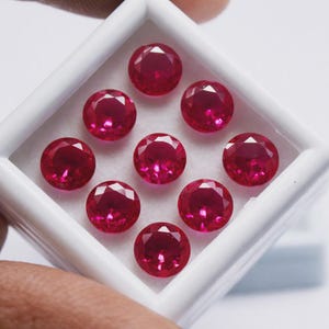 Pode incluir: Uma coleção de nove pedras preciosas redondas, facetadas e de um vermelho vibrante, dispostas numa grelha de três por três dentro de uma caixa de exposição quadrada branca. Cada pedra apresenta um corte brilhante com múltiplas facetas.