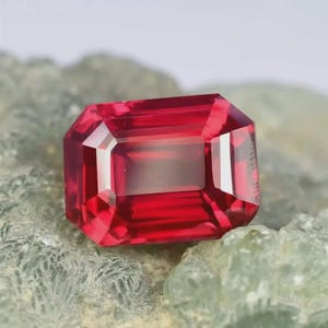 Könnte beinhalten: Ein leuchtend tiefroter Edelstein mit rechteckigem Smaragdschliff und scharfen Facetten, die das Licht reflektieren. Der Stein ist auf einer texturierten, blassgrünen Mineralmatrix platziert, was den Kontrast zwischen Rot und Grün betont.