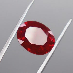 May include: A vibrant, oval-cut red gemstone held by a pair of silver-toned metal tweezers. The stone features a faceted surface that reflects light, showcasing a deep, saturated crimson hue throughout its transparent, polished crystalline structure.