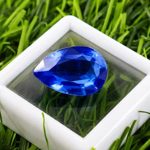 May include: A pear-shaped, faceted sapphire gemstone, deep blue in color, is displayed within a white square box. The box sits on a bed of green artificial grass, creating a striking contrast between the gem and its surroundings.