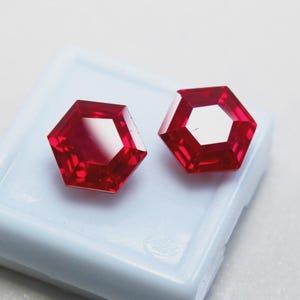 以下が含まれることがあります： 2つのファセット加工された六角形の赤い宝石。宝石は鮮やかで深い赤色で、研磨された反射面を持っています。白いプラスチックトレイにセットされており、その形と色を際立たせています。