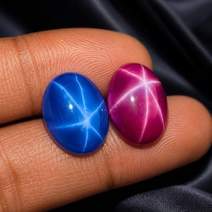 Puede incluir: Dos piedras preciosas cabujón de forma ovalada, una de color azul vibrante y otra de color magenta intenso, cada una con un efecto de estrella de seis rayos conocido como asterismo. Las piedras tienen una superficie lisa y pulida.