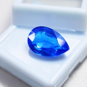 May include: A vibrant, pear-shaped, faceted blue gemstone. The gem is displayed in a white plastic jewelry box. The gemstone's facets reflect light, creating bright highlights. The overall appearance is a deep, saturated blue.