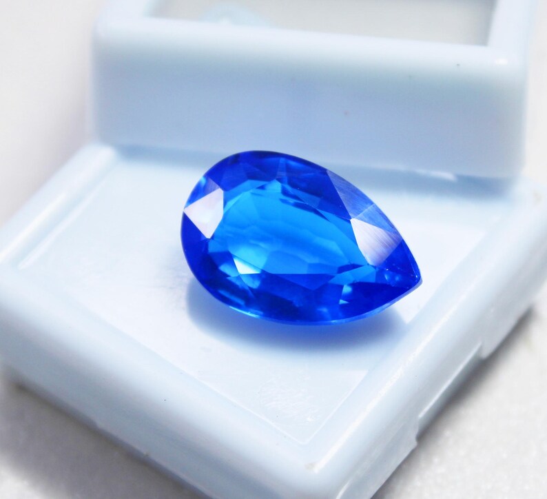May include: A vibrant, pear-shaped, faceted blue gemstone. The gem is displayed in a white plastic jewelry box. The gemstone's facets reflect light, creating bright highlights. The overall appearance is a deep, saturated blue.