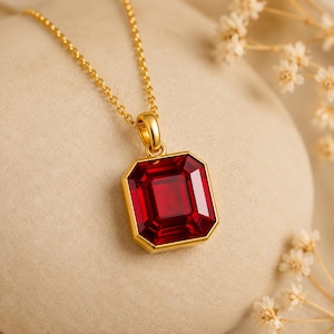 May include: A gold necklace with a large, rectangular, faceted red gemstone pendant. The pendant is set in a gold frame and hangs from a delicate gold chain. The gemstone has a deep, rich red colour. The necklace is displayed on a neutral background.