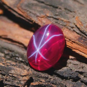 Puede incluir: Una gema ovalada pulida de color rojo rubí, con un patrón de estrella de seis rayos. La estrella está formada por líneas blancas que se cruzan. La gema se encuentra sobre un fondo de madera marrón oscuro texturizado.