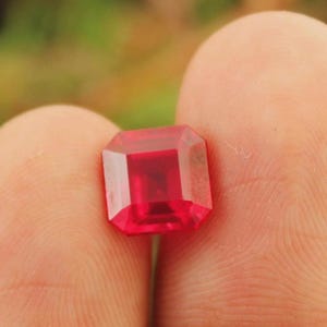 Puede incluir: Una gema roja intensa, tallada en forma rectangular y facetada. La gema se sostiene entre dos dedos, mostrando su claridad y color vibrante. Las facetas de la gema reflejan la luz, creando un efecto brillante. El fondo está borroso.