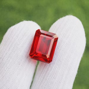 Könnte beinhalten: Ein leuchtender, rechteckiger Edelstein im Smaragdschliff mit tiefrotem Farbton und facettierten Oberflächen, die das Licht reflektieren. Der Stein weist eine hohe Reinheit und eine gesättigte karmesinrote Farbe mit präzisen geometrischen Schliffen auf.