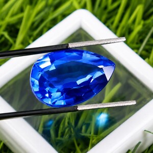 May include: A faceted, pear-shaped, deep blue gemstone is held by black tweezers against a white square frame. The gem's facets reflect light, creating a brilliant display. The background is blurred green grass.