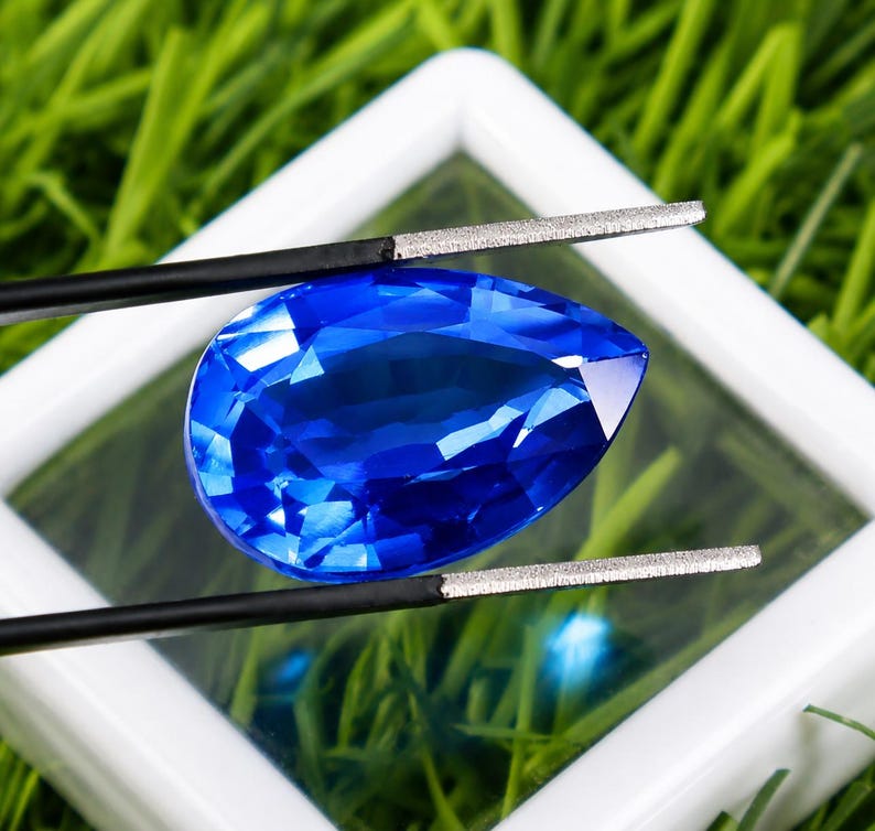 May include: A faceted, pear-shaped, deep blue gemstone is held by black tweezers against a white square frame. The gem's facets reflect light, creating a brilliant display. The background is blurred green grass.