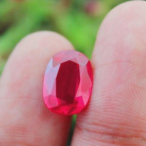 May include: A faceted, oval-cut ruby gemstone. The gem is a deep, vibrant red colour, with a polished surface that reflects light. The ruby is held between fingers, with a blurred green background.