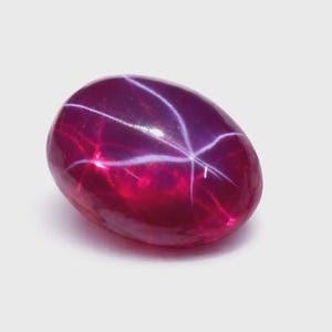 May include: An oval-shaped, polished gemstone in a deep red colour, displaying a six-rayed star effect. The gem's surface reflects light, creating a starburst pattern. The gemstone is set against a white background.