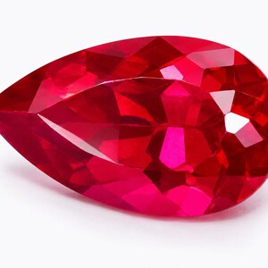 Op de afbeelding: Een peervormige, gefacetteerde edelsteen met een levendige, dieprode kleur. De steen heeft talloze geometrische facetten die het licht weerkaatsen, wat zorgt voor een schitterende en fonkelende uitstraling over het gladde, gepolijste oppervlak.