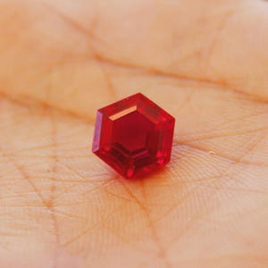 May include: A faceted, hexagonal, deep red gemstone. The gem is set against a warm, skin-toned background, highlighting its vibrant colour and geometric shape. The gem's facets catch the light, creating a sparkling effect.