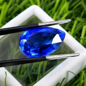 May include: A pear-shaped, brilliant blue gemstone is held by black tweezers against a white background. The gem is faceted, reflecting light. The background is a bed of green grass.