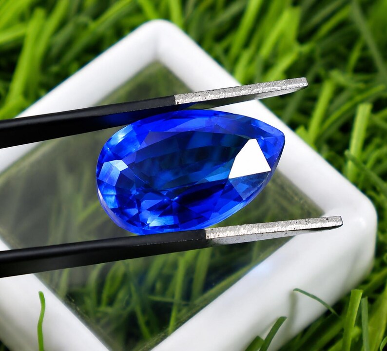 May include: A pear-shaped, brilliant blue gemstone is held by black tweezers against a white background. The gem is faceted, reflecting light. The background is a bed of green grass.
