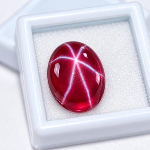 Könnte beinhalten: Ein ovaler, tiefroter Sternrubin-Edelstein mit einem ausgeprägten sechsstrahligen Asterismus-Effekt auf seiner glatten, polierten Cabochon-Oberfläche. Der Edelstein befindet sich in einer quadratischen weißen Schmuckschatulle.