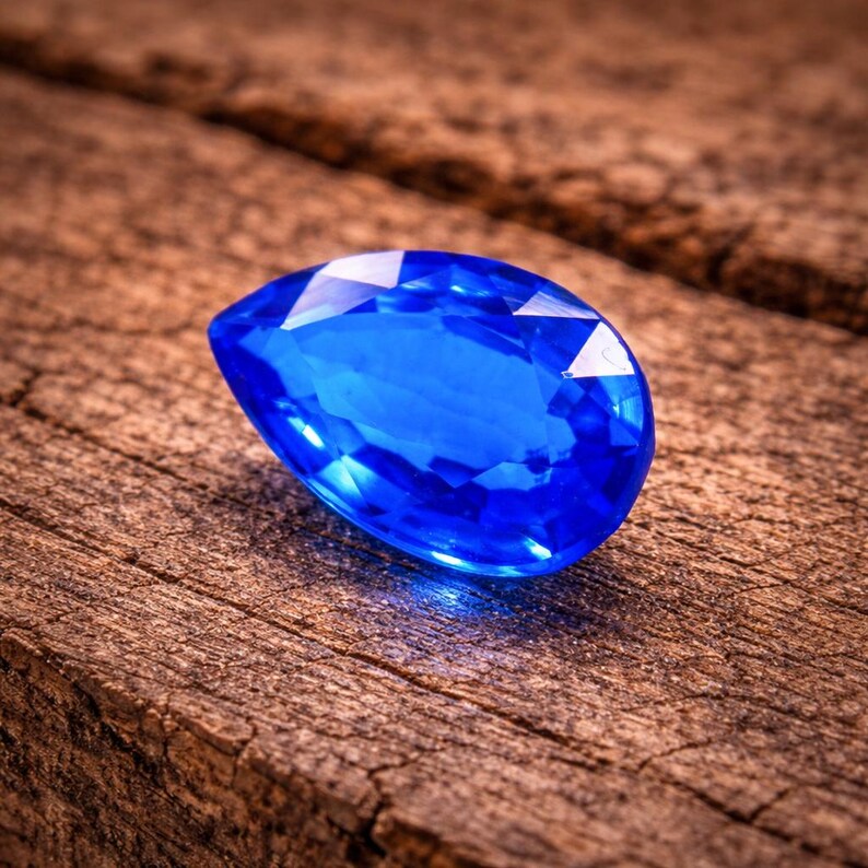 May include: A faceted, pear-shaped, brilliant blue gemstone rests on a weathered, brown wooden surface. The gem's facets reflect light, creating bright highlights. The wood grain adds a rustic contrast to the gem's polished surface.