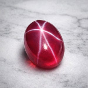 Puede incluir: Una gema de rubí pulida, de forma ovalada, con un efecto de estrella de seis rayos. La gema es de color rojo intenso con líneas blancas que irradian desde un punto central. La piedra está sobre una superficie de mármol blanco y gris.