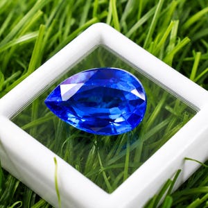 May include: A pear-shaped, faceted sapphire gemstone, displayed within a clear case with a white frame. The sapphire is a deep, vibrant blue, set against a backdrop of green grass. The gemstone's facets reflect light, creating a sparkling effect.