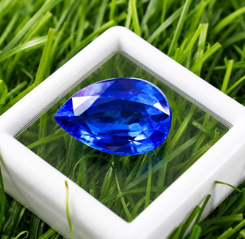 May include: A pear-shaped, faceted sapphire gemstone, displayed within a clear case with a white frame. The sapphire is a deep, vibrant blue, set against a backdrop of green grass. The gemstone's facets reflect light, creating a sparkling effect.