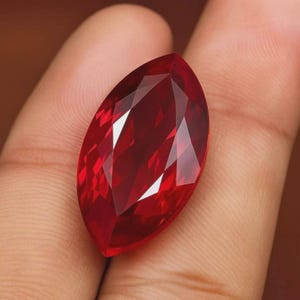 Pode incluir: Uma pedra preciosa de corte marquise com uma cor vermelha profunda e vibrante e uma superfície facetada que reflete a luz. A pedra apresenta uma forma oval pontiaguda com facetas geométricas simétricas que criam uma aparência brilhante.