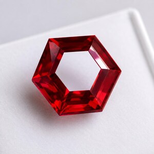 Può includere: Una gemma rosso vibrante tagliata in forma esagonale con un centro cavo. La pietra presenta molteplici sfaccettature che riflettono la luce, creando profonde tonalità cremisi e bordi geometrici netti su tutta la superficie lucida.