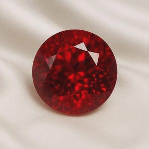 Può includere: Una gemma rotonda sfaccettata di un colore rosso intenso e vibrante. La pietra presenta un taglio preciso e simmetrico con molteplici sfaccettature triangolari e a forma di aquilone che creano un complesso motivo di riflessione.