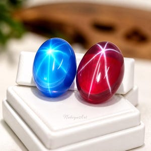 May include: Two oval cabochon gemstones, one vibrant blue and one deep red, each displaying a distinct six-rayed star effect on their polished surfaces. The stones are positioned side-by-side on a white display stand with the text NabiyaArt visible.
