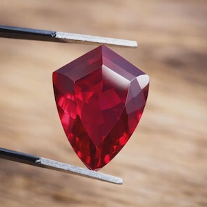 May include: A faceted, shield-shaped ruby gemstone held by tweezers. The ruby is a deep, saturated red colour with multiple facets that reflect light. The background is a blurred wooden surface.