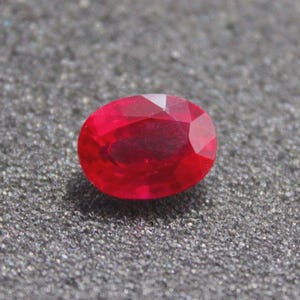 Könnte beinhalten: Ein facettierter, oval geformter Rubin-Edelstein. Der Edelstein hat eine leuchtend rote Farbe, mit mehreren Facetten, die das Licht einfangen. Der Rubin liegt auf einer strukturierten, grauen Oberfläche.