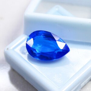 May include: A vibrant, pear-shaped, faceted blue gemstone. The gem is displayed in a light blue plastic case, showcasing its brilliant color and clarity. The gemstone's facets reflect light, creating a sparkling effect.