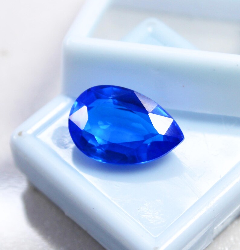 May include: A vibrant, pear-shaped, faceted blue gemstone. The gem is displayed in a light blue plastic case, showcasing its brilliant color and clarity. The gemstone's facets reflect light, creating a sparkling effect.