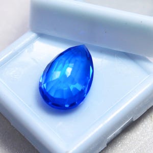 May include: A faceted, teardrop-shaped, brilliant blue gemstone. The gem is set against a light blue background, showcasing its clarity and color. The gem's facets reflect light, creating a sparkling effect. This gemstone is likely for jewelry making.