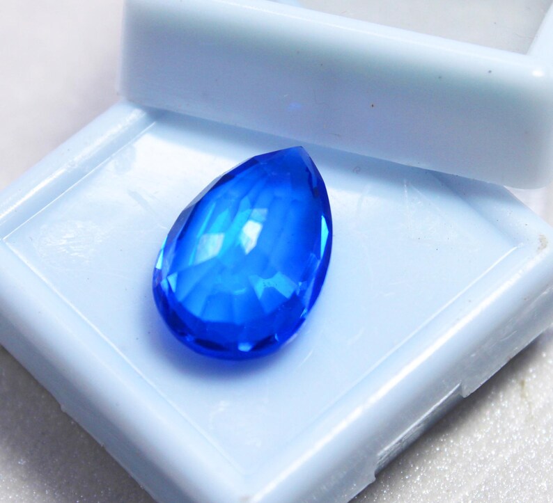 May include: A faceted, teardrop-shaped, brilliant blue gemstone. The gem is set against a light blue background, showcasing its clarity and color. The gem's facets reflect light, creating a sparkling effect. This gemstone is likely for jewelry making.