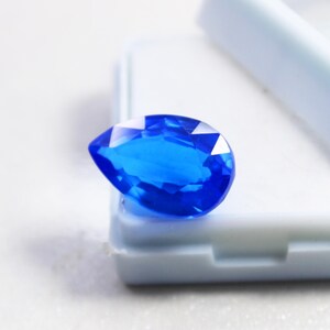 May include: A faceted, pear-shaped, brilliant blue gemstone. The gem is set against a white background, showcasing its clarity and vibrant color. The cut of the stone allows light to reflect, creating a sparkling effect. This gemstone is ideal for jewelry making.