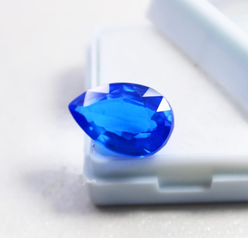 May include: A faceted, pear-shaped, brilliant blue gemstone. The gem is set against a white background, showcasing its clarity and vibrant color. The cut of the stone allows light to reflect, creating a sparkling effect. This gemstone is ideal for jewelry making.