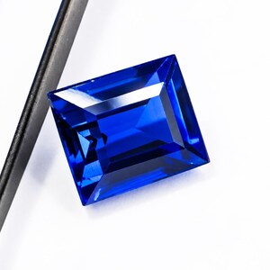Può includere: Una pietra preziosa blu rettangolare sfaccettata con una tonalità zaffiro profonda e vibrante. La pietra presenta tagli geometrici precisi che creano bordi netti e riflessi di luce interni. È posizionata accanto a un sottile strumento metallico scuro.