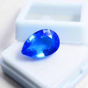 May include: A brilliant, pear-shaped, faceted blue gemstone rests in an open, light blue jewelry box. The gemstone's facets reflect light, showcasing its vibrant color and clarity. The box is white inside.