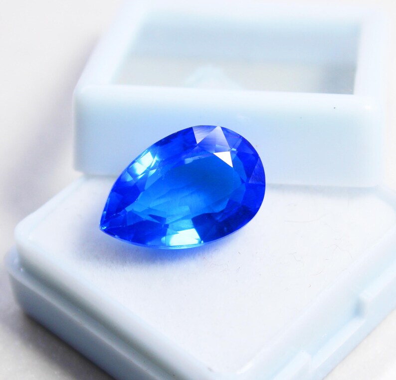 May include: A brilliant, pear-shaped, faceted blue gemstone rests in an open, light blue jewelry box. The gemstone's facets reflect light, showcasing its vibrant color and clarity. The box is white inside.