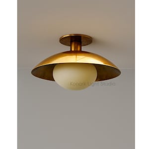 May include: A brass-colored ceiling light fixture with a rounded shade and a white globe-shaped light. The fixture is mounted on a white ceiling. The text "Konark Light Studio" is visible on the light.