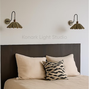 May include: Two wall sconces with a dark bronze finish and a pleated, olive-green shade are mounted on a white wall above a bed. The bed has a dark brown headboard, beige pillows, and a decorative pillow with a black and tan zebra pattern. The text "Konark Light Studio" is visible.