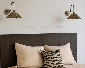 May include: Two wall sconces with a dark bronze finish and a pleated, olive-green shade are mounted on a white wall above a bed. The bed has a dark brown headboard, beige pillows, and a decorative pillow with a black and tan zebra pattern. The text "Konark Light Studio" is visible.