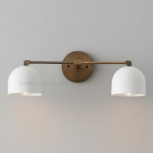 Può includere: Applique da parete a due luci con una piastra posteriore rotonda in ottone anticato e un braccio orizzontale. Due paralumi bianchi a forma di cupola sono attaccati al braccio. L'applique è progettata per uso interno e ha un'estetica moderna. Il testo "Konark Light Studio" è visibile.