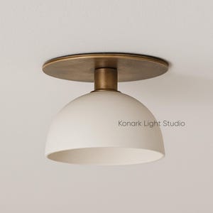 May include: A modern ceiling light fixture with a white, dome-shaped shade and a brass-colored mounting plate and stem. The fixture is mounted on a white ceiling. The text "Konark Light Studio" is printed on the shade.