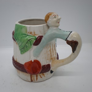 May include: A white ceramic mug with a brown, red, and green design. The mug has a handle shaped like a person with a blue shirt and a brown hat.