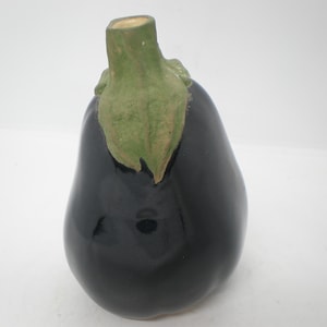 May include: A ceramic eggplant figurine with a glossy, dark purple body. The stem and leaf are green and tan. The eggplant is a decorative item, possibly for a kitchen or dining area.