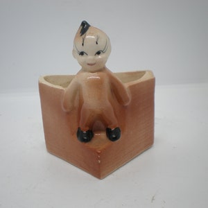 Puede incluir: Un macetero de cerámica con forma de casa con una pequeña figura de persona de dibujos animados de pie en la puerta. La figura tiene el pelo negro, un cuerpo marrón y zapatos negros. El macetero es de color marrón claro.