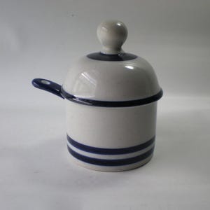 May include: A white ceramic sugar bowl with a lid and handle. The bowl has two horizontal blue stripes and a blue rim. The lid has a blue rim and a small, round knob. The handle is blue.