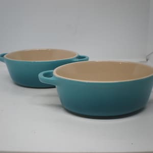 May include: Two teal ceramic bowls with tan interiors and small handles. The bowls are oval-shaped and sit on a white surface. The bowls are likely for serving or baking.